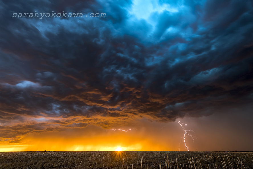 A nighttime, tornadic mezocyclone lightning storm shoots bolt of electricity to the ground and lights up the field and dirt road in Tornado Alley. A large lightning strike at dusk in an open plain framed against a deep, dark orange sunset and stormy skies.  A large lightning strike at dusk in an open plain framed against a deep, dark orange sunset and stormy skies.